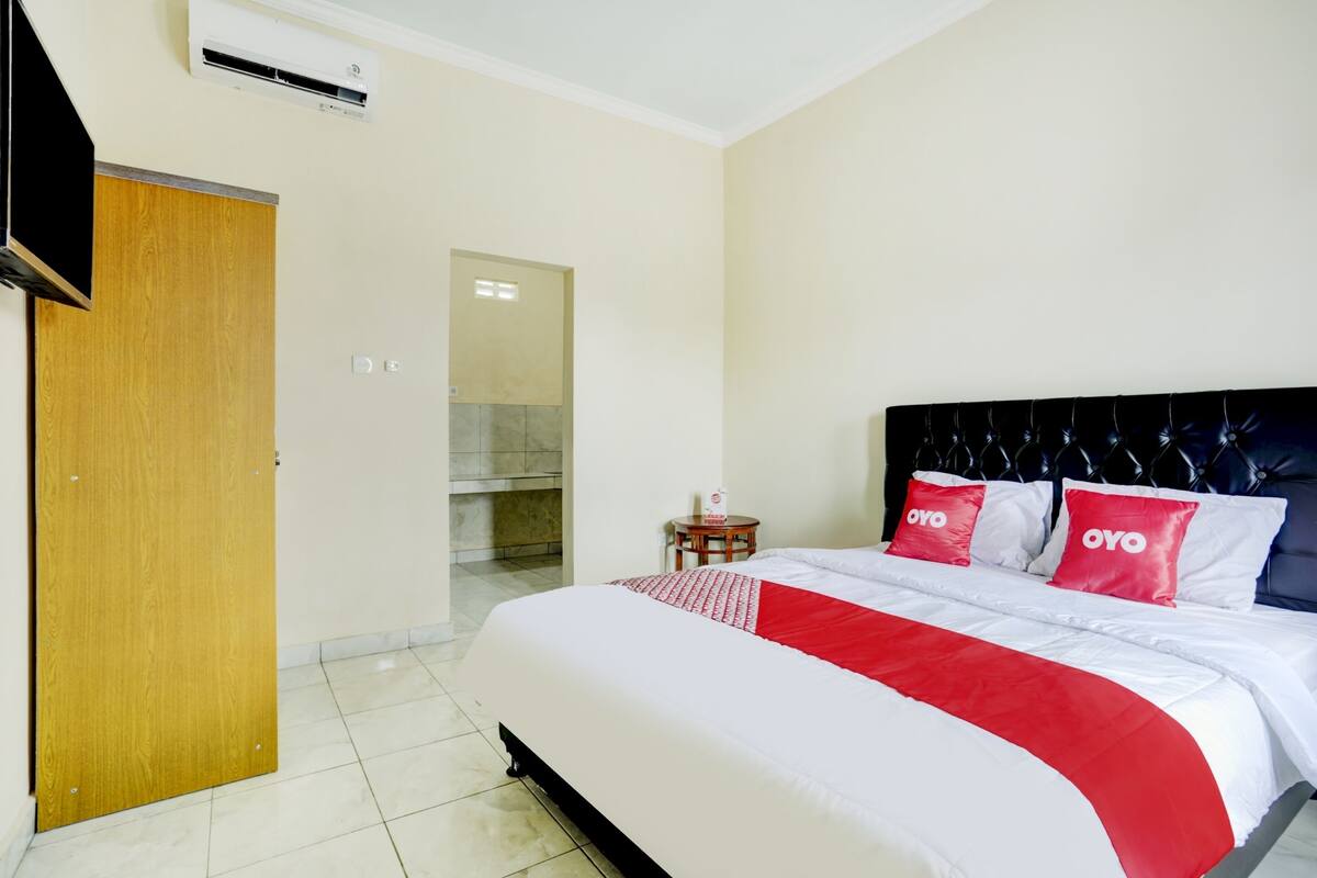 A hotel room with a bed, a wooden door, an air conditioning unit, and a small round table with a bottle.