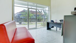 A room with a red sofa, a desk, and a large sliding glass door leading to a courtyard with plants and a building. - Hotel O Pondok Dukuh (Nusa Dua)