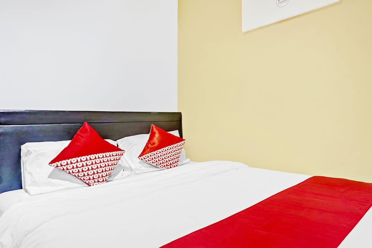 Cama com roupa de cama branca e almofadas vermelhas, cabeceira escura e parede amarela com uma moldura.