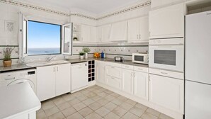 Cocina moderna con armarios blancos, horno empotrado, microondas y lavavajillas. Una ventana ofrece vistas al mar.