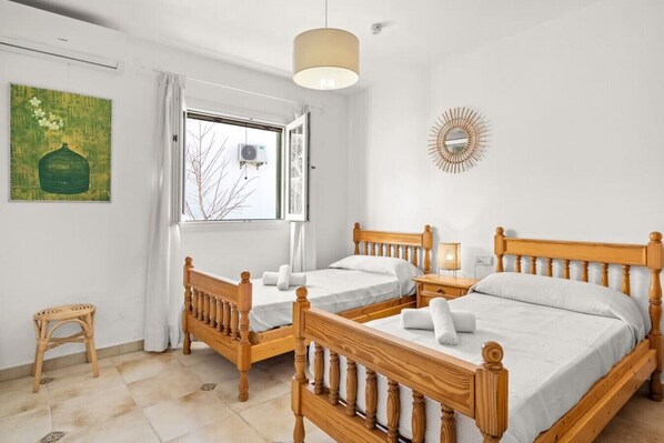 A hotel room with two wooden beds, a small wooden stool, a window with curtains, a wall-mounted air conditioner, and a decorative mirror. - Villa Atalis, Stunning sea views (San Jaime Mediterráneo)