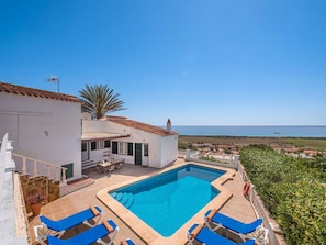 A pool area with lounge chairs, a white building with a red-tiled roof, and a view of the sea.