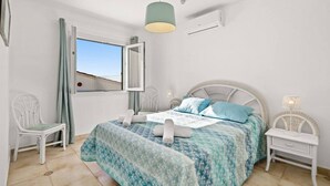 Un dormitorio con cama, dos sillas, una mesita de noche, una ventana con cortinas y un equipo de aire acondicionado.