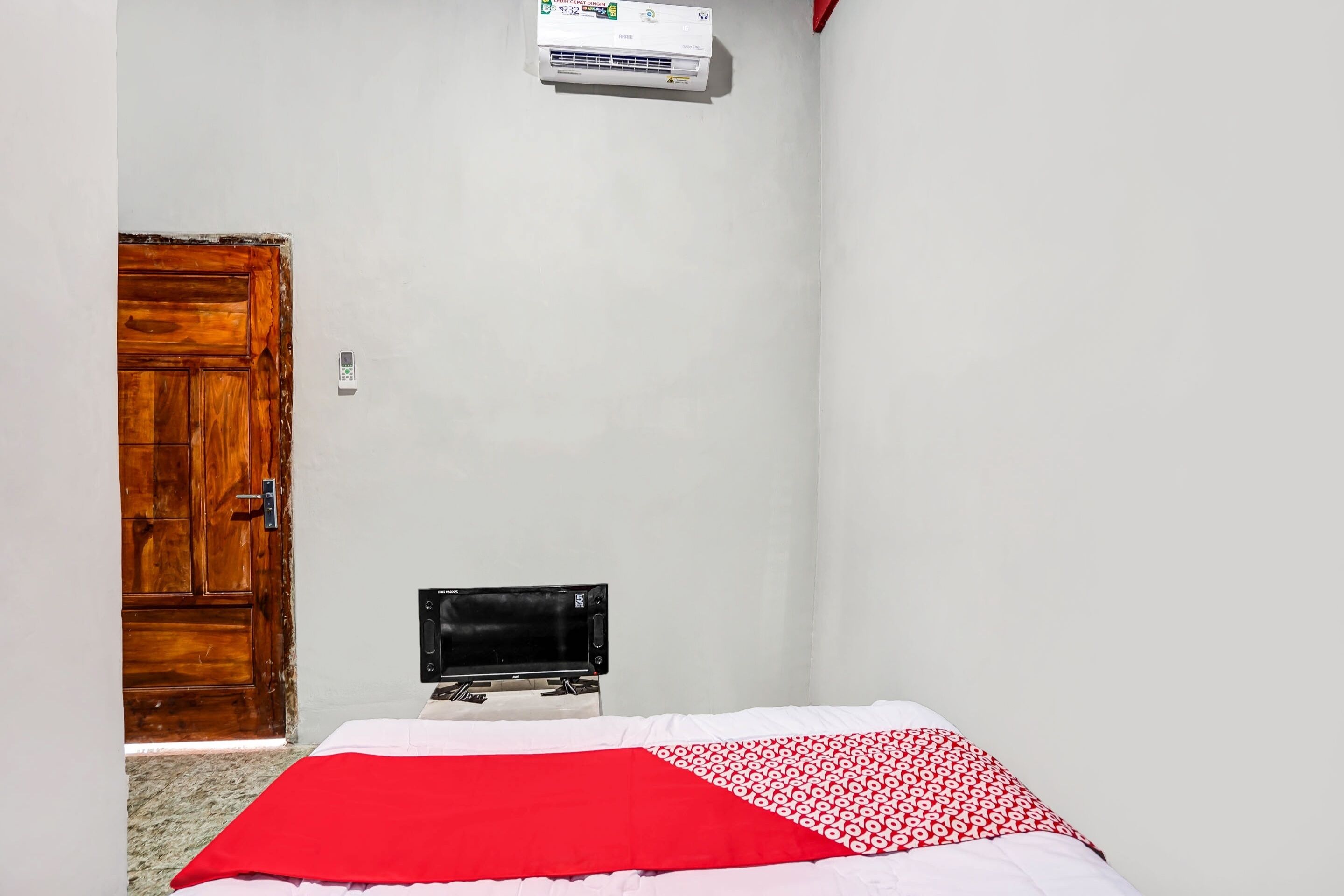 A room with a wooden door, a bed with a red and white patterned blanket, a wall-mounted air conditioner, and a television on a stand.