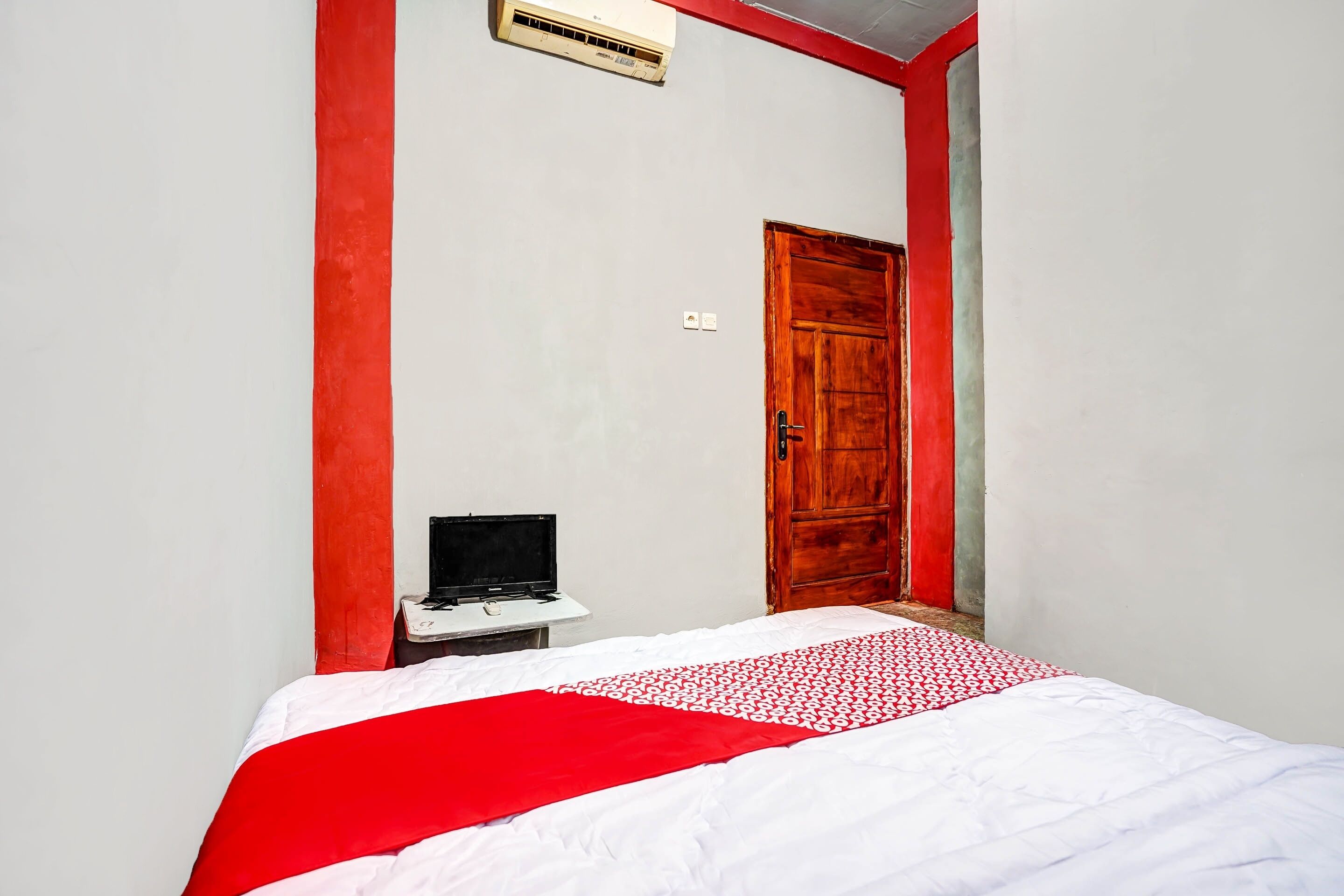 Quarto com cama, porta de madeira, uma pequena mesa com um laptop e um edredom vermelho e branco.