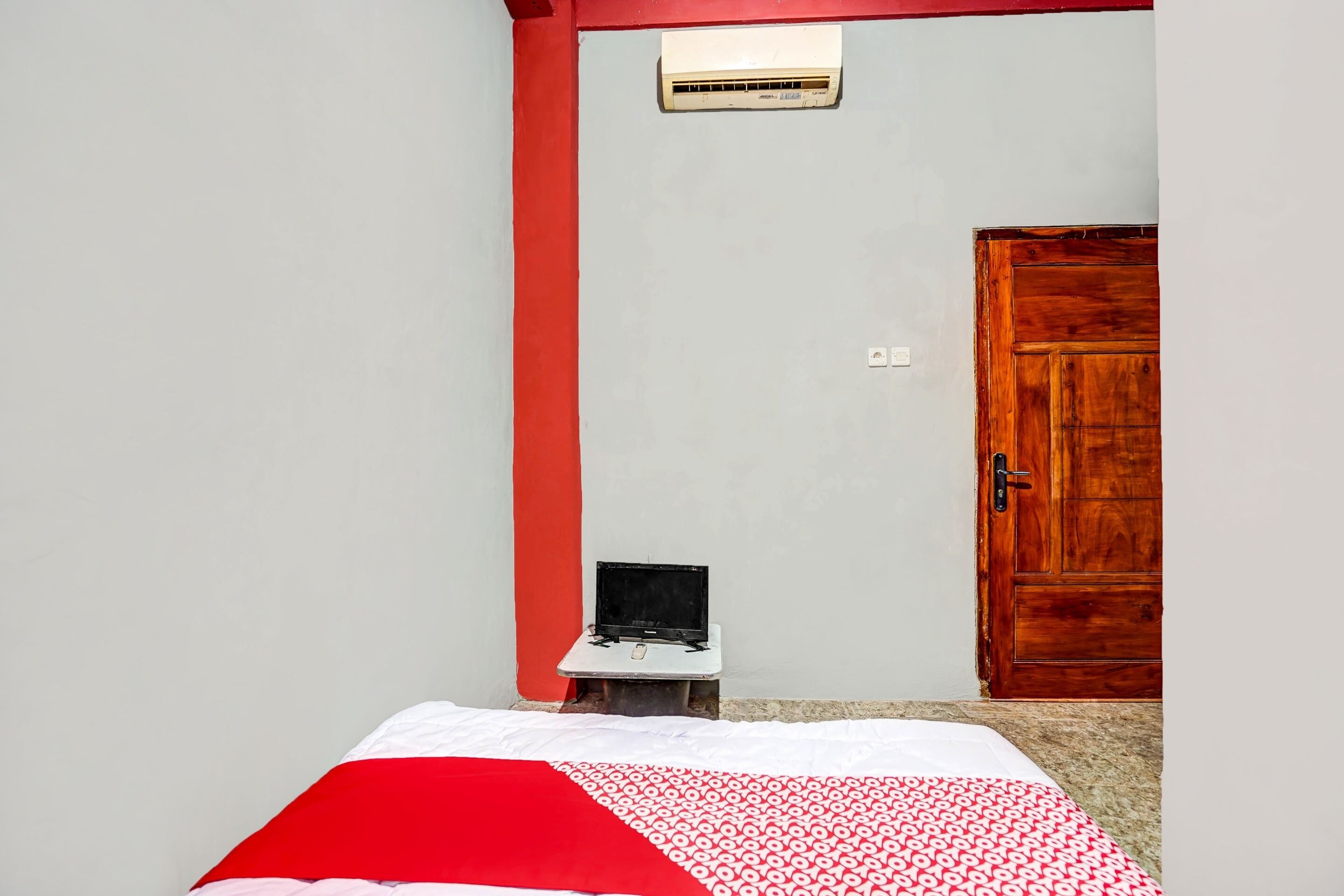 A bedroom with a bed, a small table with a laptop, a wooden door, and a red-framed window.