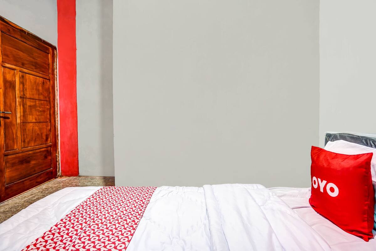 Een kamer met een houten deur, een bed met een deken met wit en rode patronen, en een rood kussen met de tekst 'OYO'.