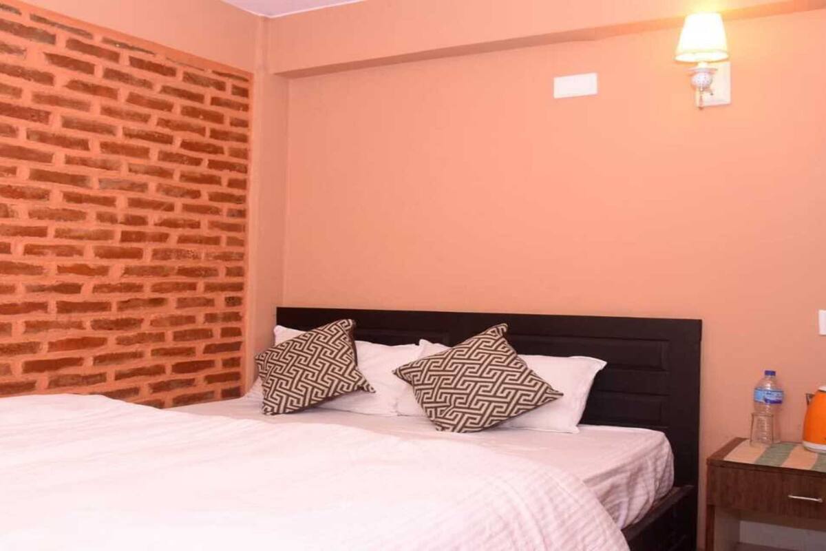 Une chambre d’hôtel avec un mur décoré d’un motif en briques, un lit agrémenté d’oreillers à motifs, une table de chevet avec une bouilloire et une bouteille d’eau, et une lampe fixée au mur.