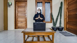 Een persoon zittend aan een houten tafel met een laptop, voor een houten deur en een raam met gordijnen. Twee potten met cactussen staan naast de deur.