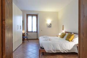 Une chambre à coucher comprenant un lit, deux oreillers, une table de chevet, une fenêtre avec des rideaux, une chaise et une armoire.