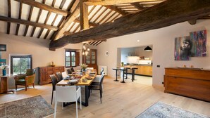 Une salle à manger spacieuse avec un plafond en bois, une grande table à manger dressée pour accueillir plusieurs personnes, et une vue sur la cuisine en arrière-plan.
