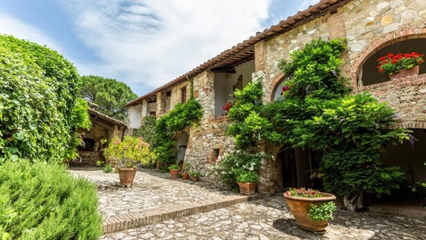 Une cour pavée de pierres, agrémentée de plantes en pot, et un bâtiment rustique doté d’arches au niveau des fenêtres et d’un toit recouvert de tuiles.