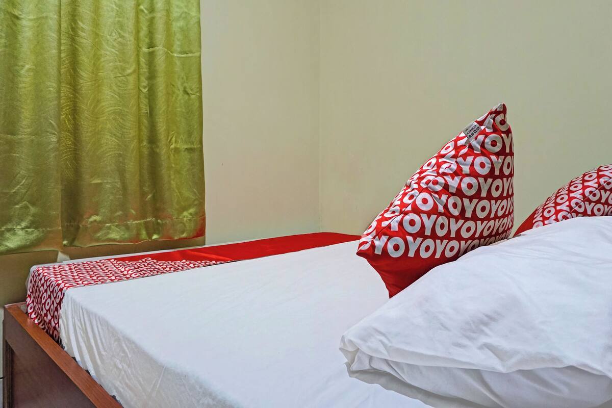 A bed with white bedding and red and white patterned pillows, a green curtain, and a plain wall.