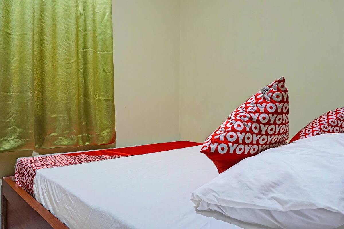 A hotel room with a bed, red and white patterned pillows, a white bedspread, and a green curtain.