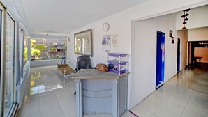 A hallway with a desk, chair, and shelves; a clock on the wall; and a door with a blue frame. - Hotel O Darussalam Inn (Medan)