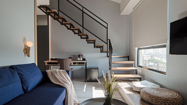 Una sala de estar moderna con un sofá azul, una mesa de centro con una planta, una escalera con baranda de malla y vistas a la ciudad a través de una ventana.