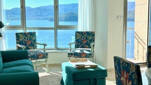Ein Zimmer mit zwei floralen Sesseln, einem türkisfarbenen Sofa und Meerblick durch große Fenster.