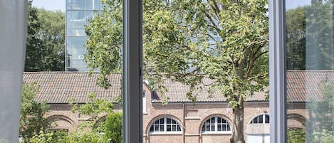 Vista desde una ventana con baranda metálica, que da a un edificio de ladrillo con ventanas arqueadas y un árbol en primer plano.