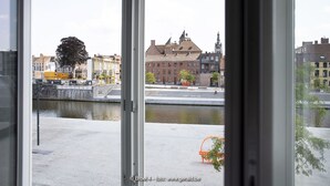 A view through a window of a building with a river in the foreground, a bridge, and buildings with distinct architectural styles in the background. - BROEL4 (Kortrijk)