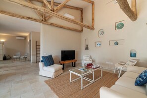 Ein modernes Wohnzimmer mit einem weißen Sofa, einem Couchtisch und einem Fernseher. An der Wand hängen dekorative Teller und es gibt einen Holzbalken an der Decke.