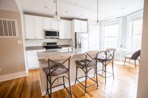 A modern kitchen with white cabinets, a central island with bar stools, and a large window providing natural light. - Lux Condo in the Heart of OTR | Walk Everywhere (Cincinnati)