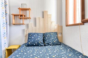 Un dormitorio con cabecera de madera, cama con colcha celeste a lunares, mesita de noche amarilla y ventana con cortina.