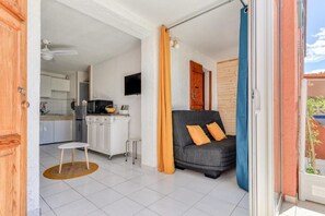 Un salon moderne avec un canapé gris, des coussins orange, une petite table blanche et un coin cuisine visible au fond.