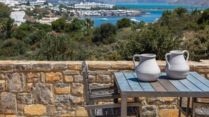 Muro de piedra con una mesa y dos jarrones blancos, con vistas a un pueblo costero con edificios y mar azul.