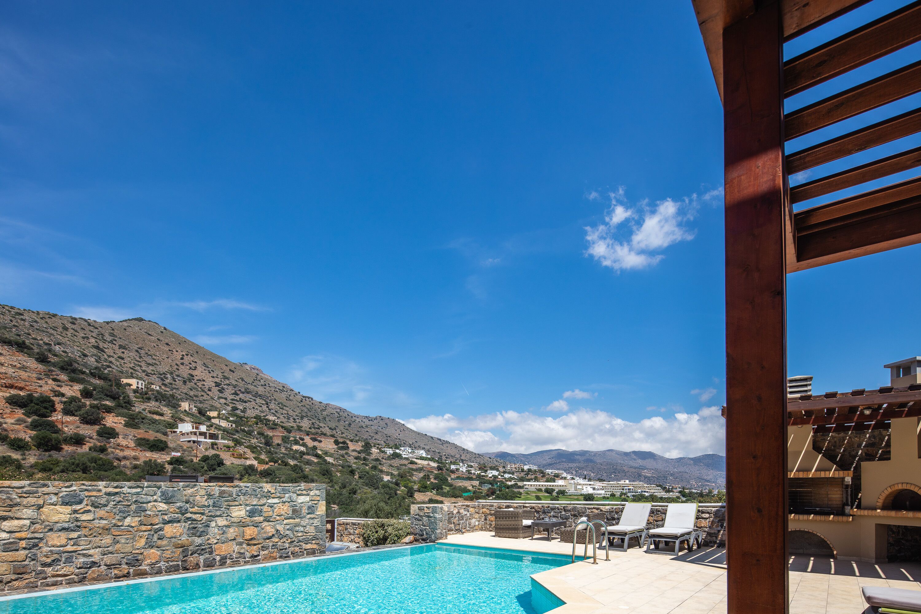 A swimming pool with a stone wall, lounge chairs, and a view of a mountainous landscape.