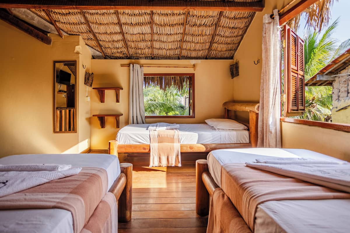 A room with three wooden beds, a thatched roof, a window with a view of greenery, and a wooden bench.