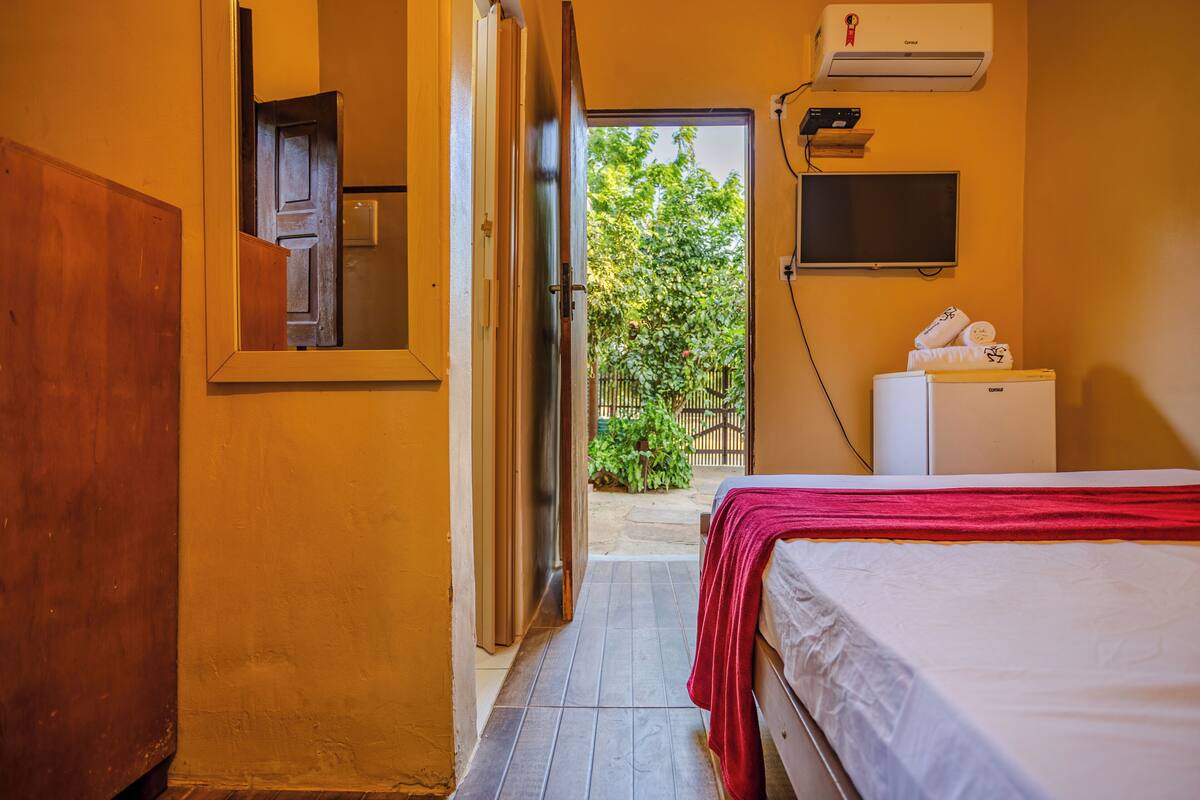 A hotel room with a bed, a small refrigerator, a TV, and an air conditioning unit.