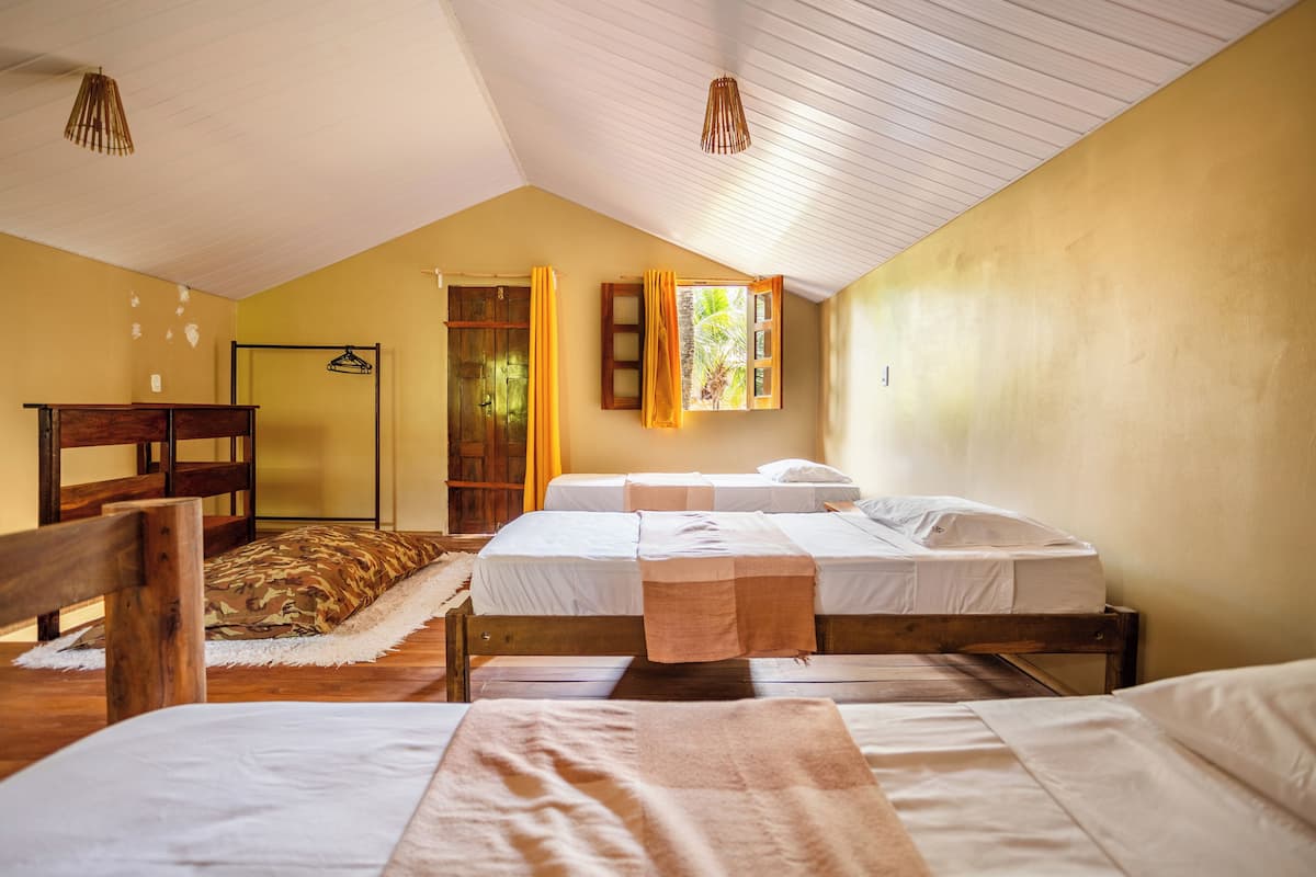 A room with two beds, wooden headboards, a wooden bench, a hanging clothes rack, and a window with curtains.