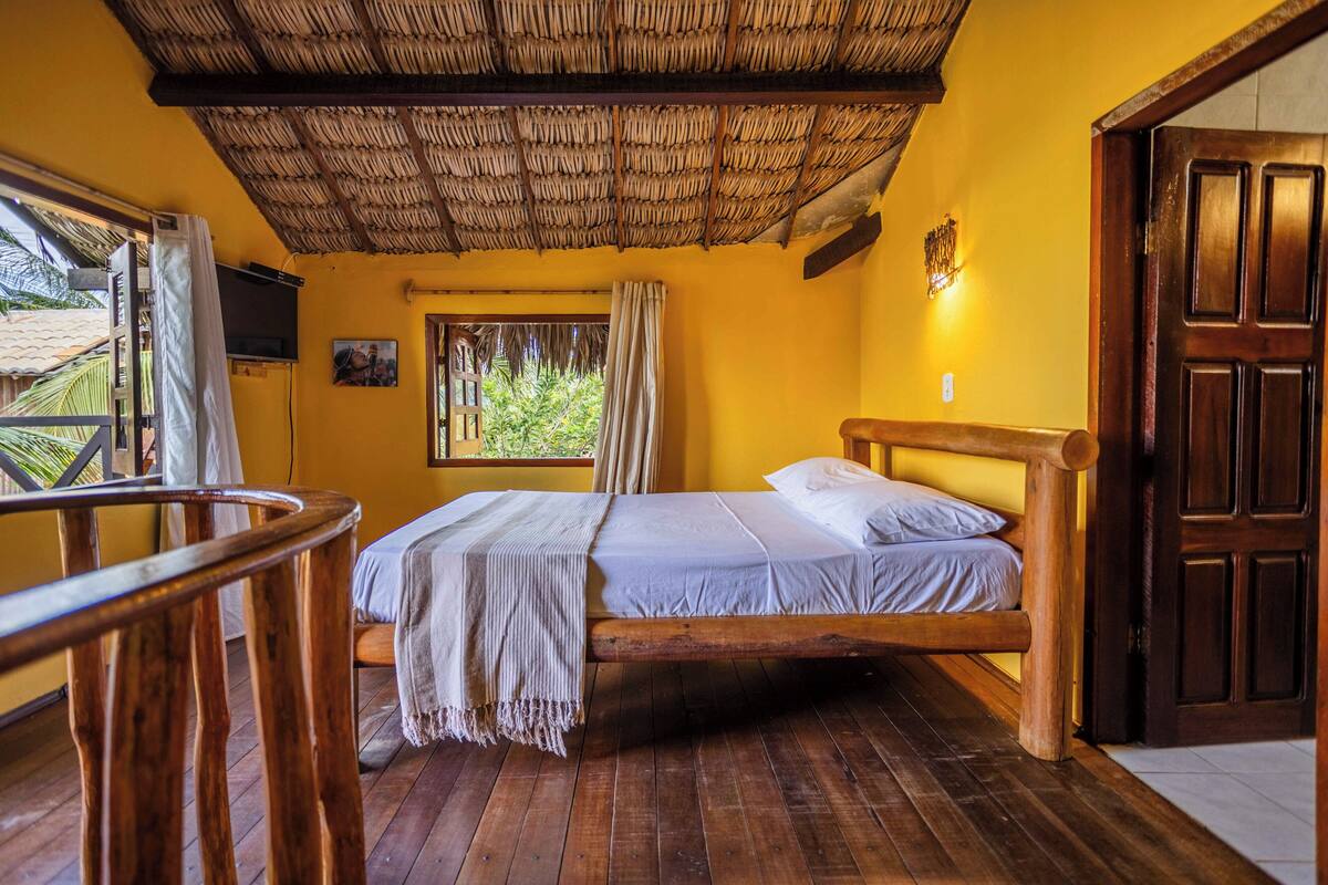 A room with a wooden bed, a window with curtains, a wooden ceiling, and a door.