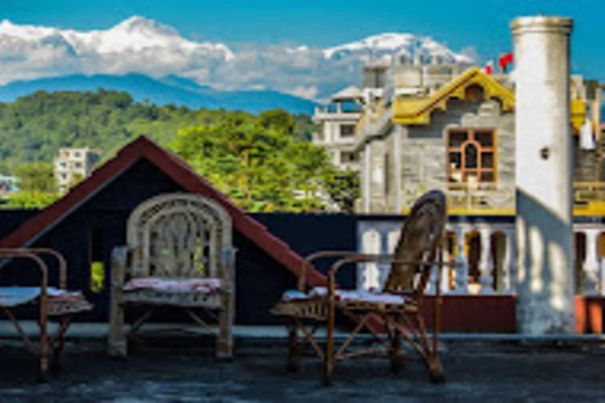 A traditional building with a sloped roof and a stone chimney, surrounded by lush greenery and a mountainous backdrop.