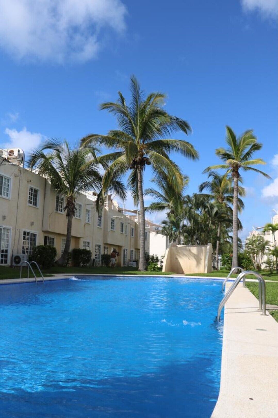 A swimming pool with clear blue water, surrounded by palm trees and a building with multiple windows.