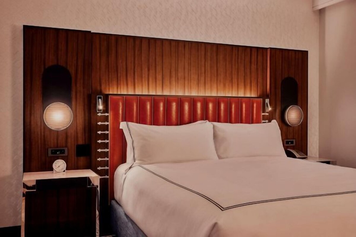 A hotel room with a wooden headboard, two bedside lamps, a nightstand with a clock, and a bed with white linens.