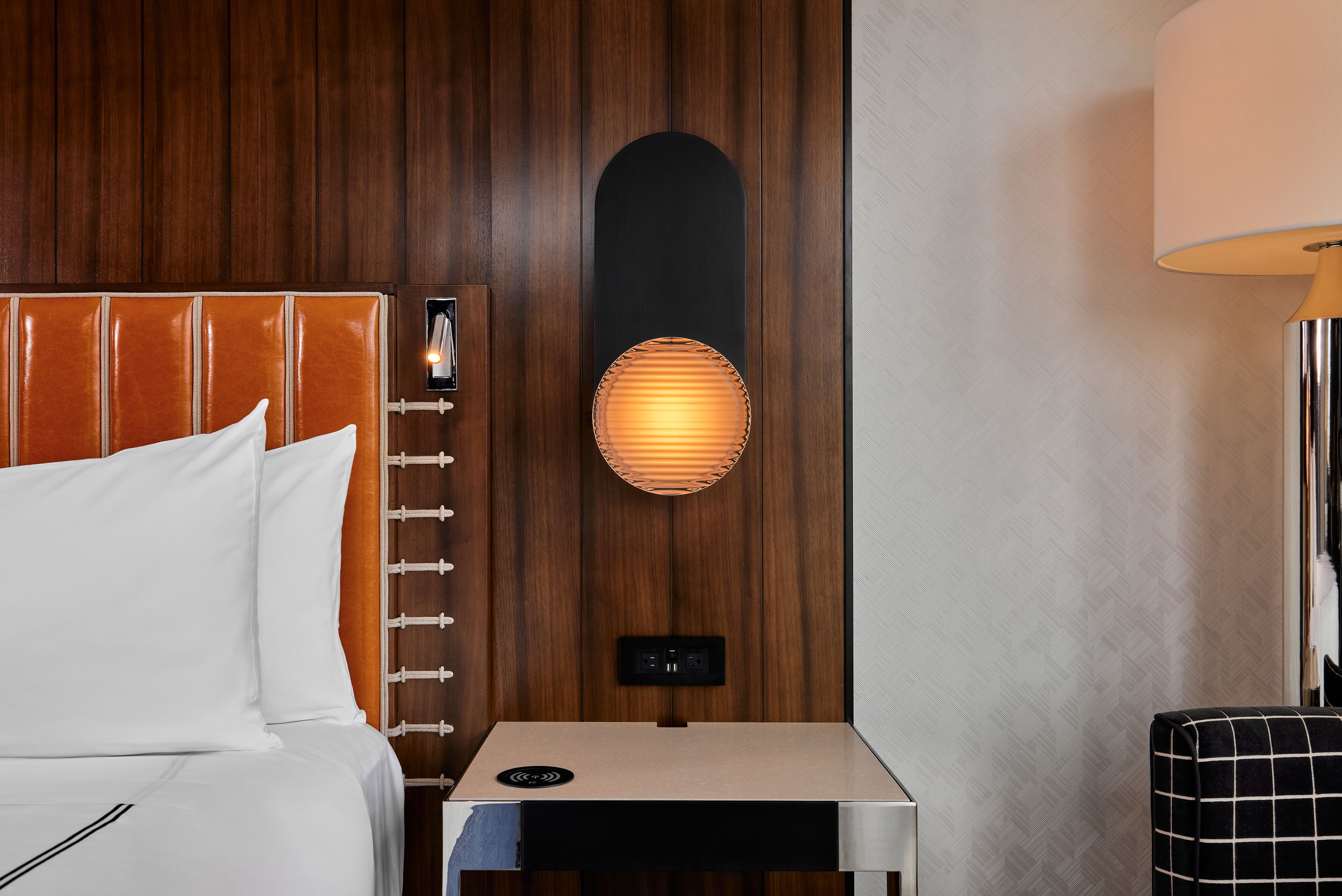 Habitación de hotel con cabecera de madera, mesita de noche con lámpara y pequeña mesa con toma de corriente.