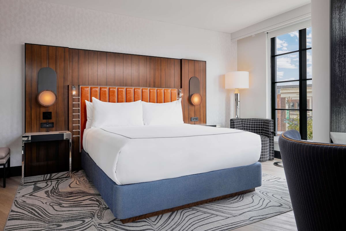A hotel room with a large bed, wooden headboard, two bedside lamps, a nightstand, a chair, and a view of the city through a window.