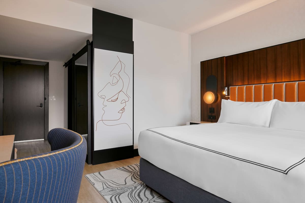 A modern hotel room with a large bed, a seating area with a chair, a wall-mounted artwork, and a wooden headboard.