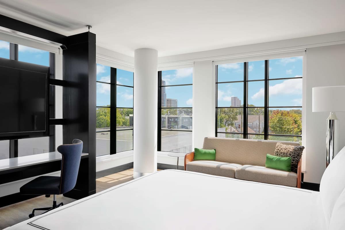 A modern hotel room with a large bed, a desk, a chair, a sofa with cushions, and large windows offering a city view.