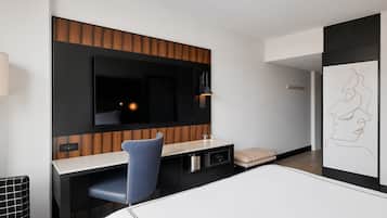A modern hotel room with a flat-screen TV mounted on a wooden panel wall, a desk with a chair, a bedside table, and a wall-mounted artwork.