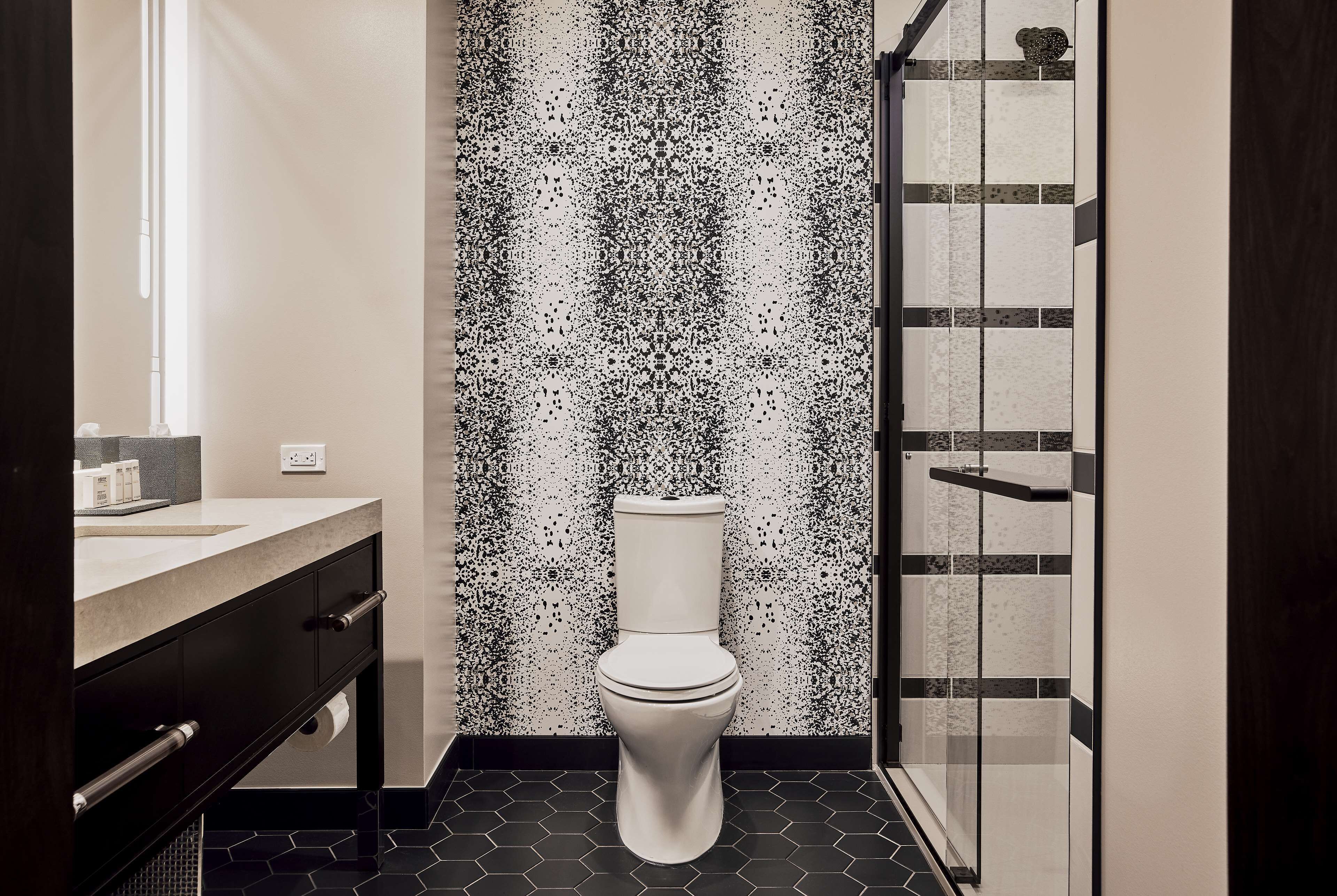 Casa de banho moderna com sanita branca, parede salpicada a preto e branco, cabine de duche em vidro e chão em mosaico escuro.