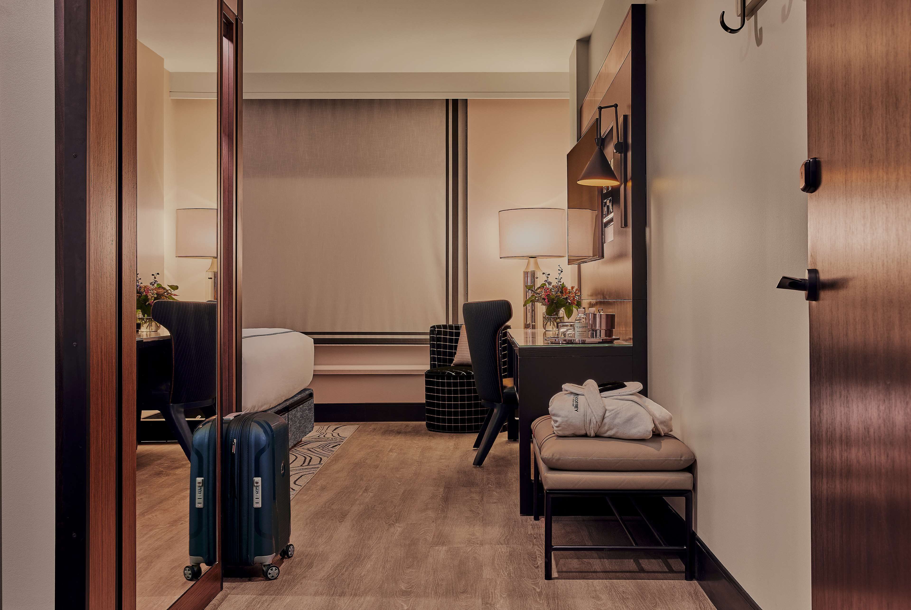Habitación de hotel con cama, escritorio, silla, sofá, lámpara, maleta y espejo.