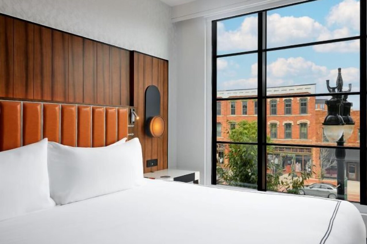 A hotel room with a large window, a wooden headboard, and a view of a cityscape.