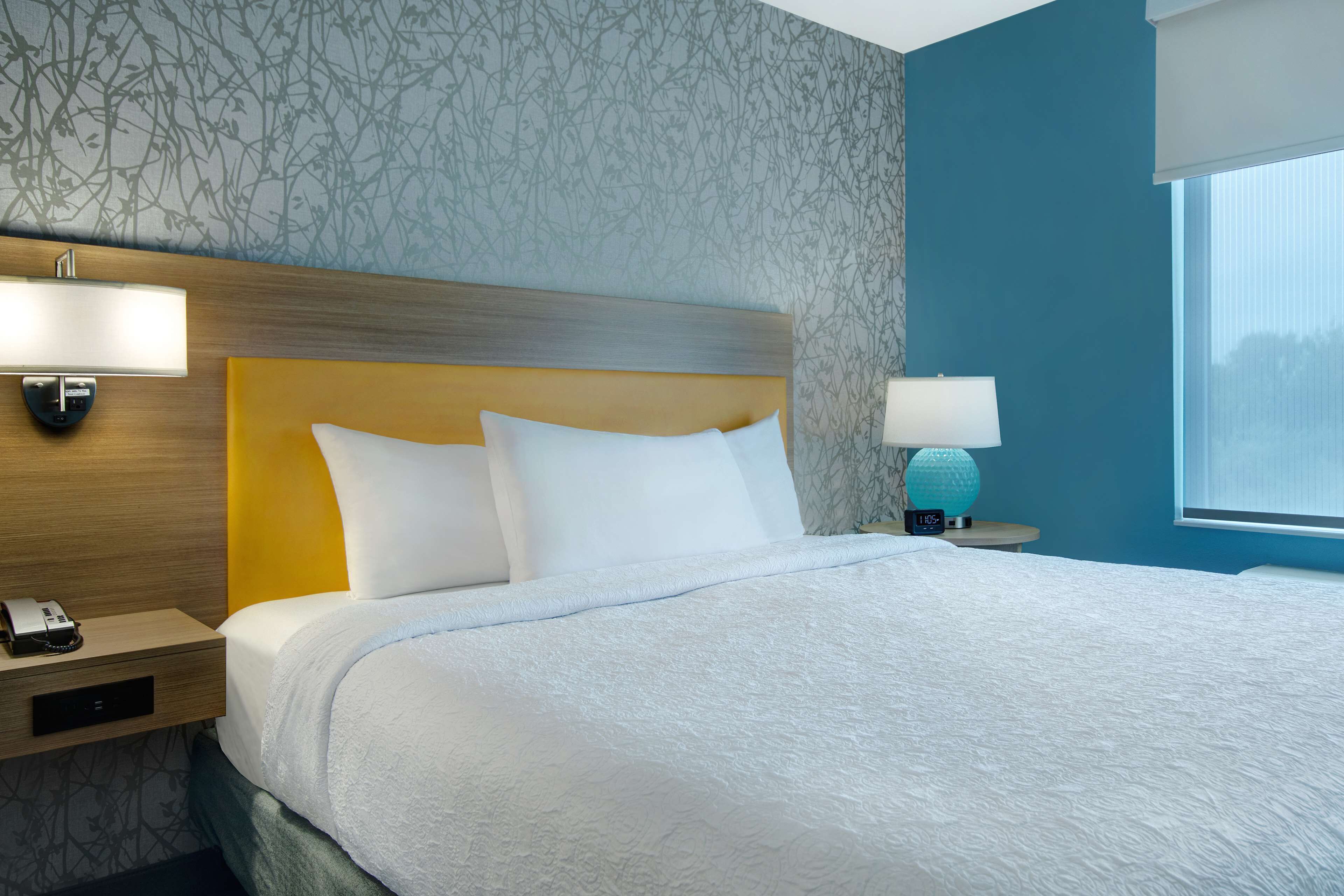 Una cama bien hecha con sábanas blancas, cabecera de madera y mesita de noche con lámpara y teléfono.