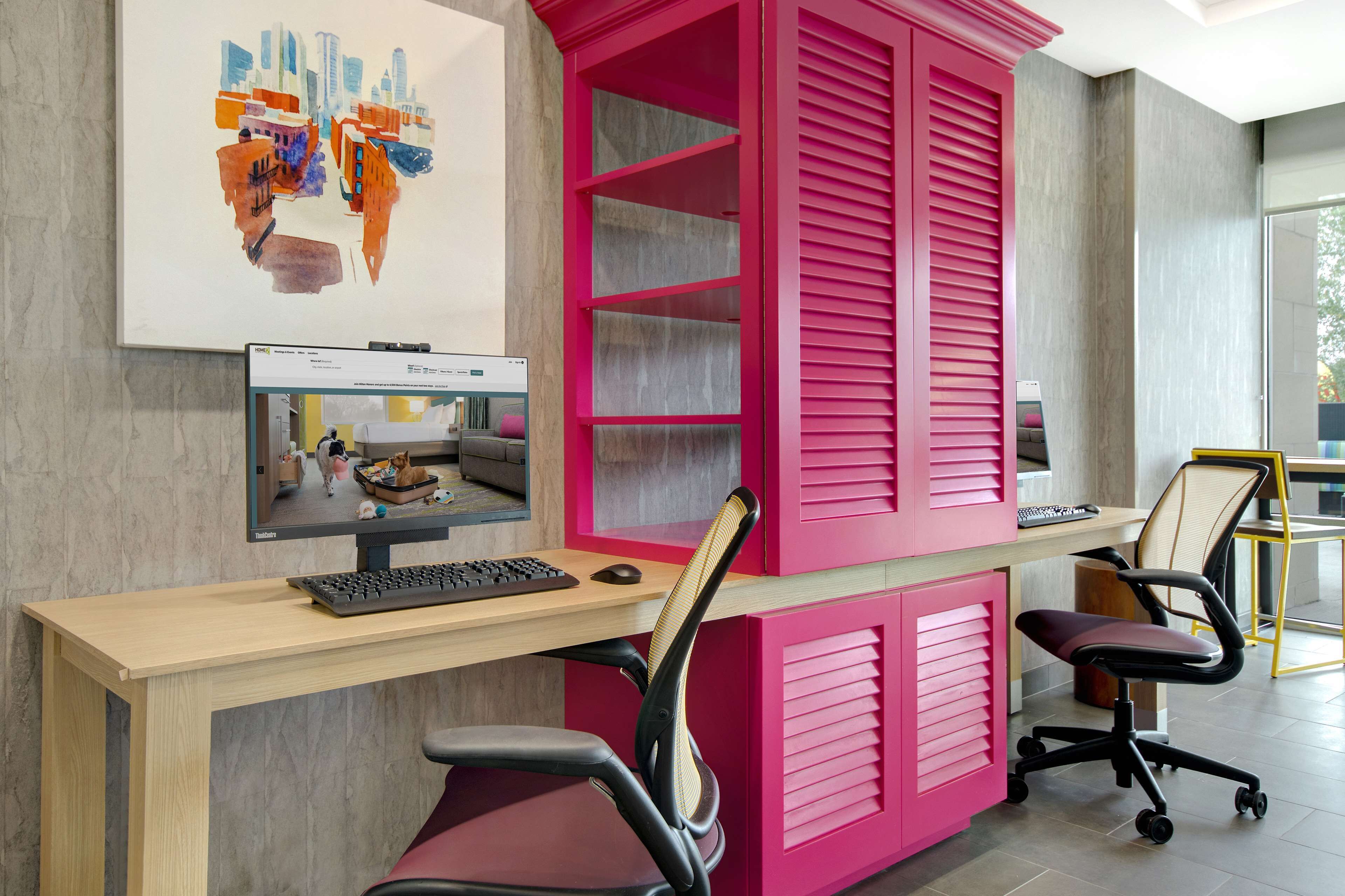 Un moderno spazio ufficio con una scrivania in legno, un mobile contenitore rosa e un monitor del computer che mostra una videochiamata.