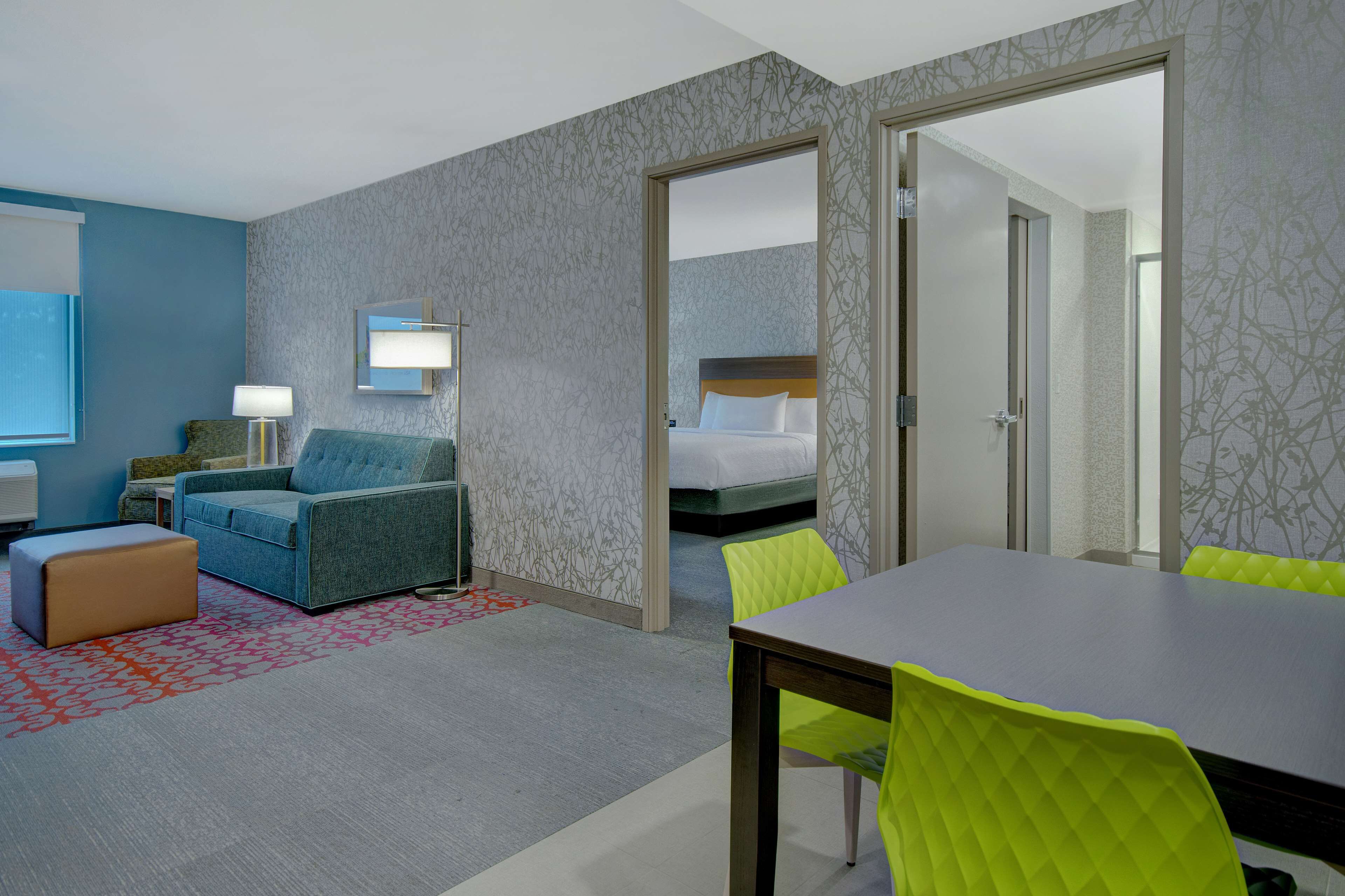 A hotel room with a blue sofa, a wooden table, and bright green chairs. There is a bed with white bedding and a mirror on the wall.
