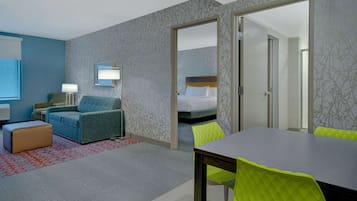 Habitación de hotel con un sofá azul, una mesa de madera y sillas verde brillante. Se ve una cama con ropa de cama blanca y un espejo en la pared.