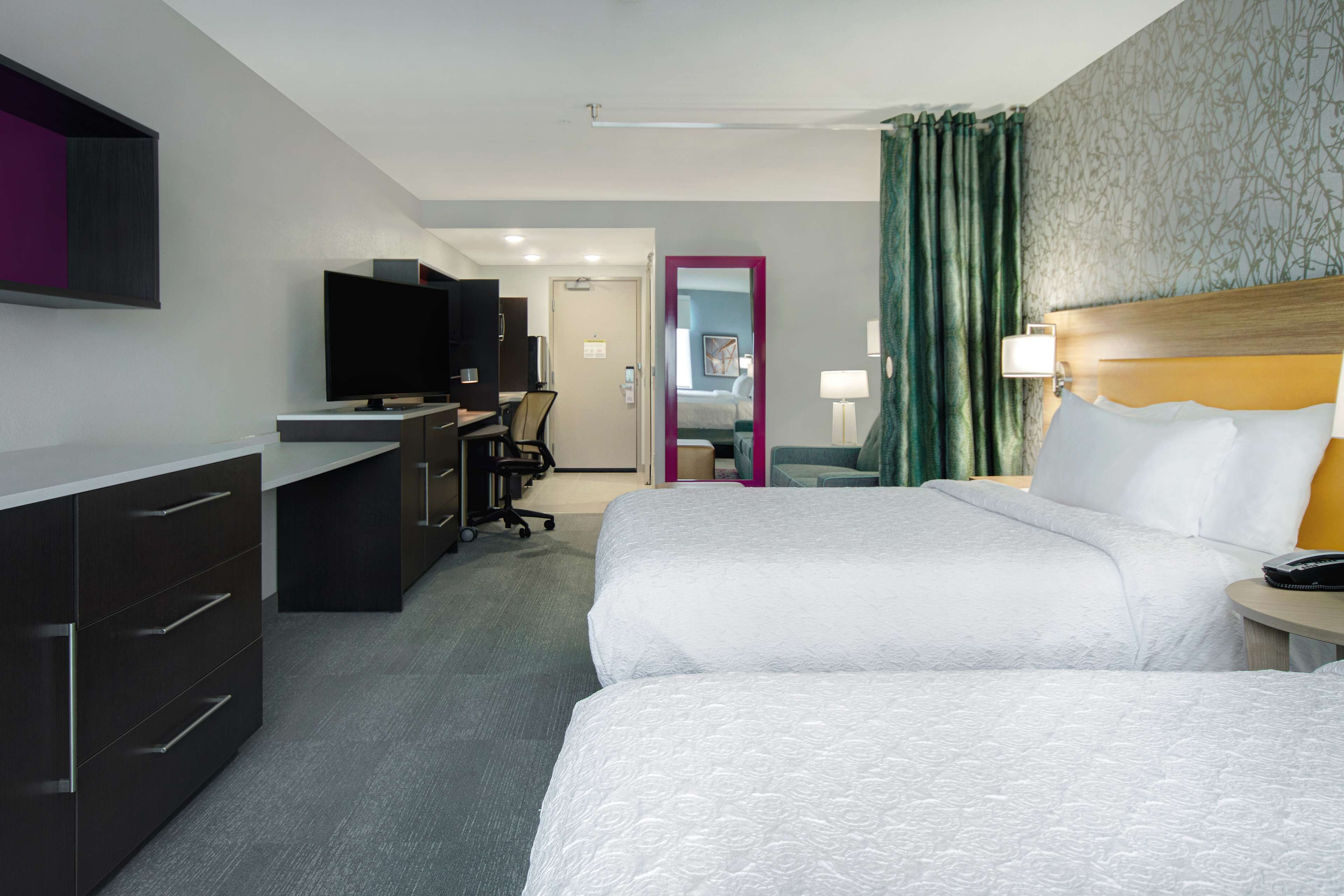 A hotel room with two beds, a desk with a flat-screen TV, a mirror, and a bathroom visible through an open door.