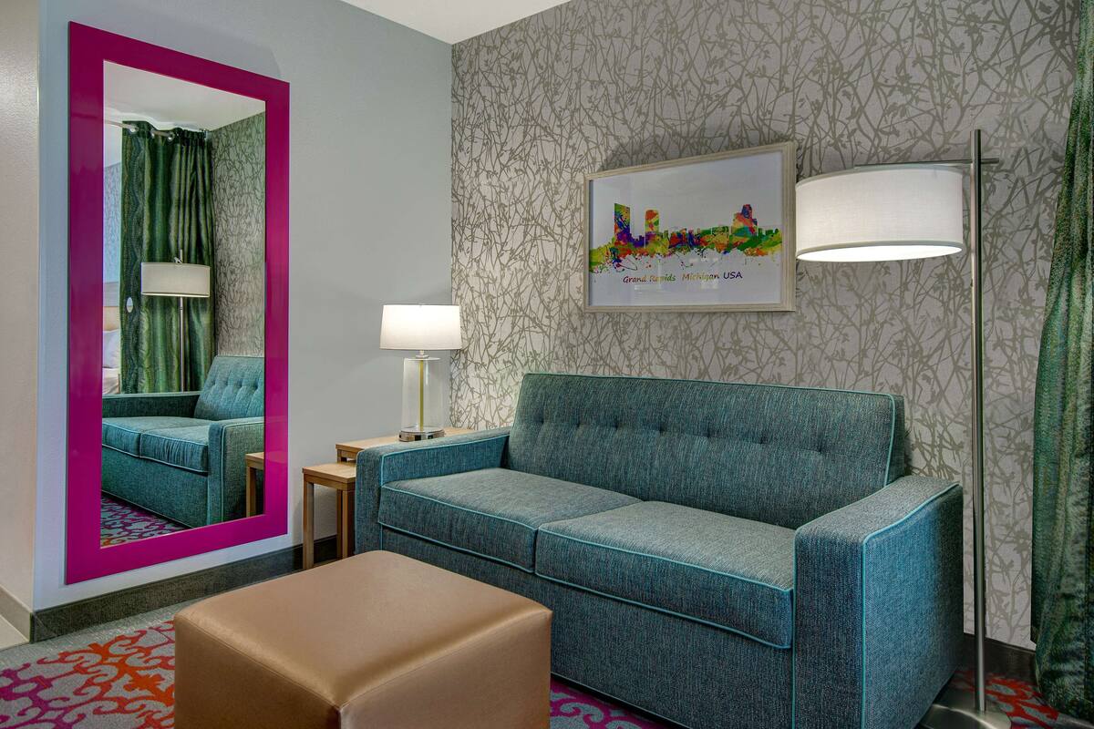 A hotel room with a teal sofa, a brown ottoman, a floor lamp, a mirror, and a framed cityscape picture on the wall.